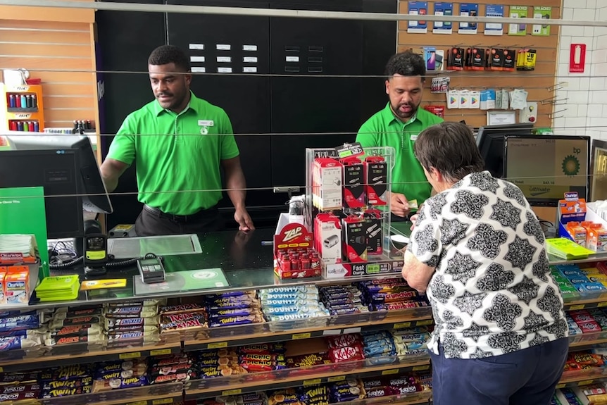 Two men standing behind a counter at a service station and serving a female customer