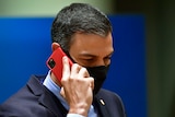 A man wearing a suit and black face mask holds an iPhone with a red case to his ear.