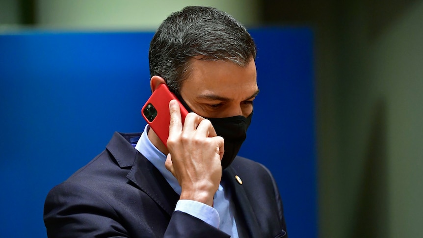 A man wearing a suit and black face mask holds an iPhone with a red case to his ear.