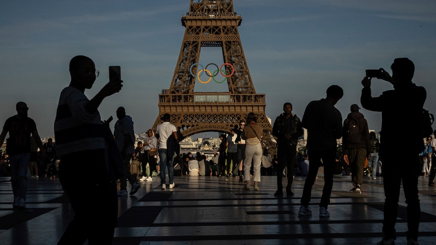 Silhouettes of people taking photos on their phones on a square in front of the Eiffel tower which has the Olympic rings on it