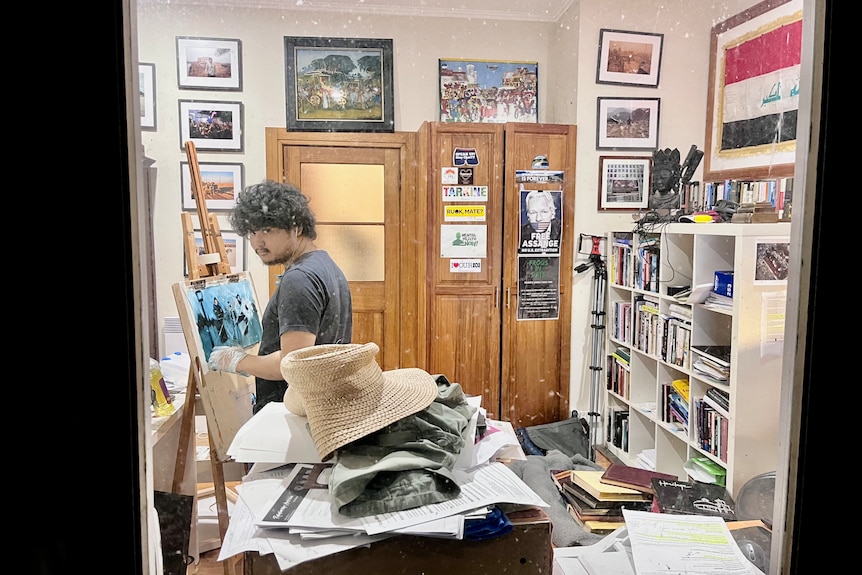 A young man with dark hair stands a a painting easel in a study. View is from an outside through the window