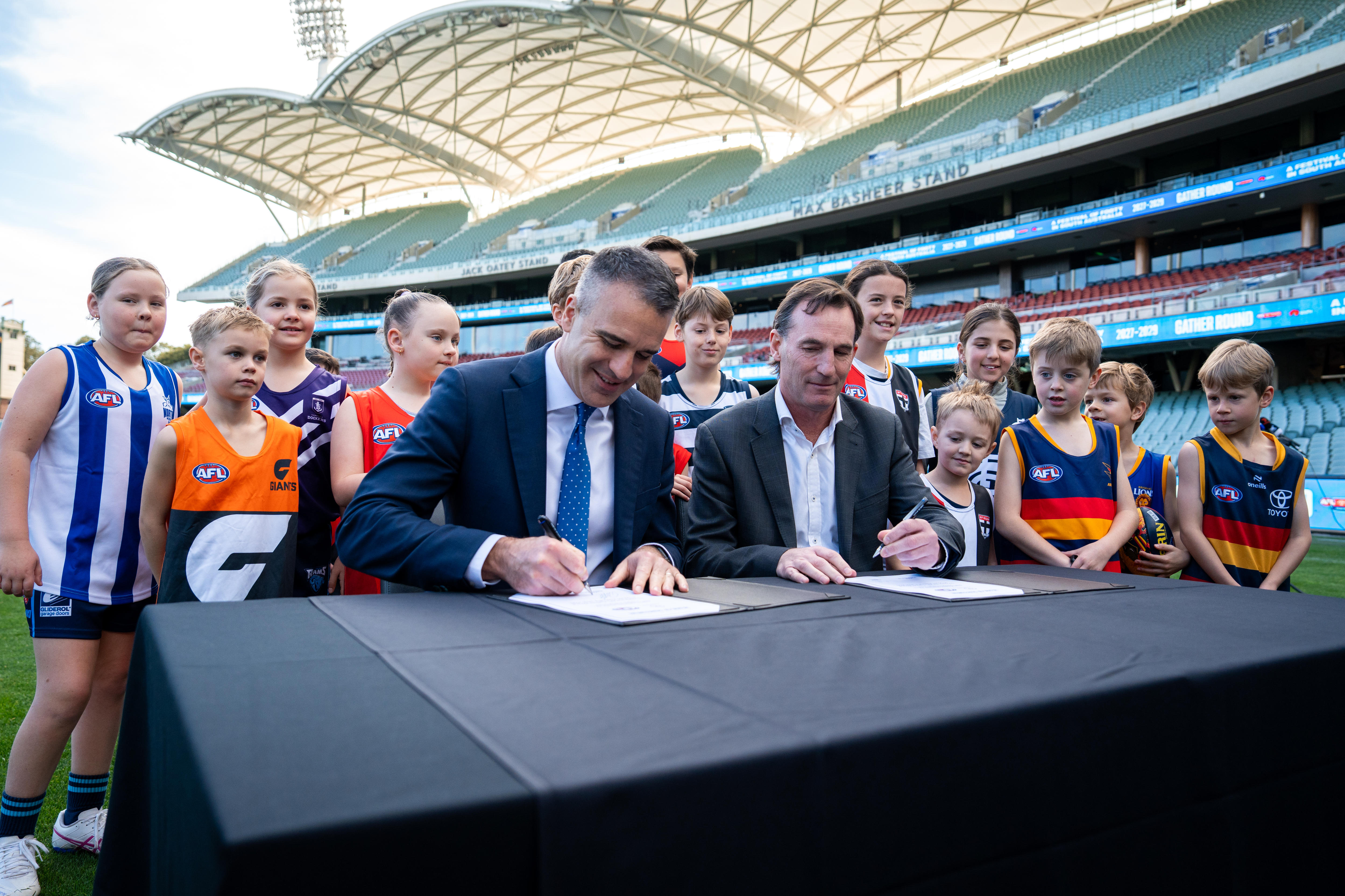 Two men sign papers on a black table while children wearing AFL jumpers look on within a stadium