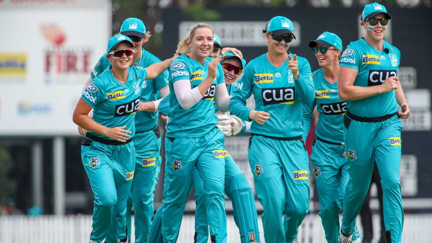 A smiling WBBL cricketer (not wearing sunglasses) jogs with her teammates after taking a wicket.