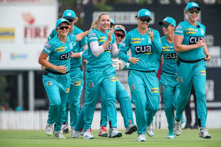 A smiling WBBL cricketer (not wearing sunglasses) jogs with her teammates after taking a wicket.