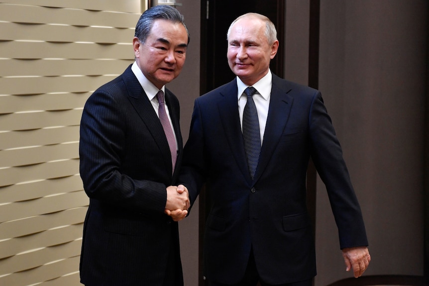 Two men in dark suits shake hands and smile as they pose for a photo in a corridor.