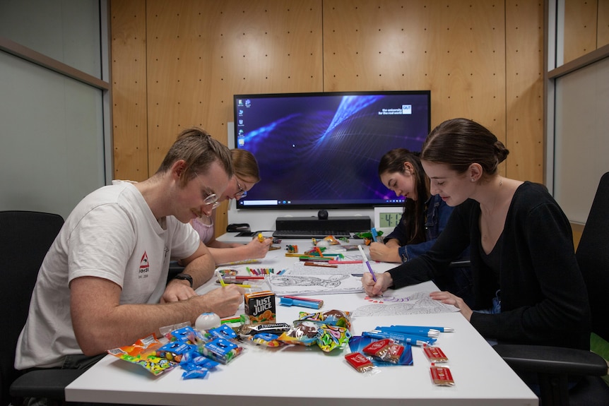 Students at a table using colour pens with snacks strewn about. 