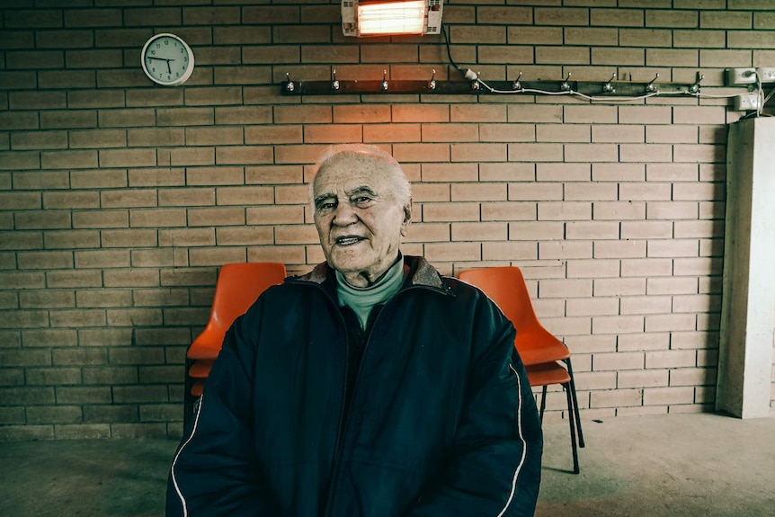 An old man in a turtleneck shirt and sports jacket sits in front of a brick wall with an orange glow from the heater above him.