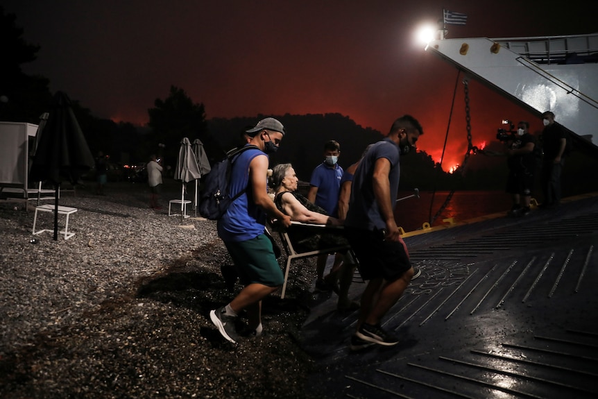 Dwóch młodszych mężczyzn pomaga starszej kobiecie pływać promem w nocy, podczas gdy w oddali płonie ogień