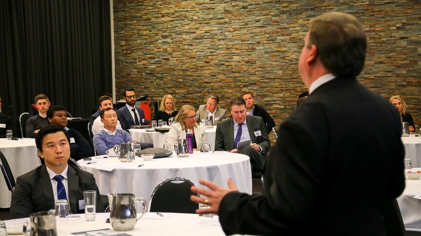 A crowd of people seated at tables listens to Peter Daly.