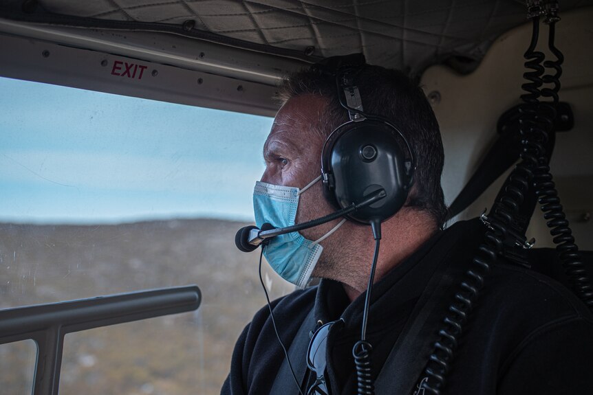 A man in a chopper, with a headset and face mask on, looks out into the mountains.