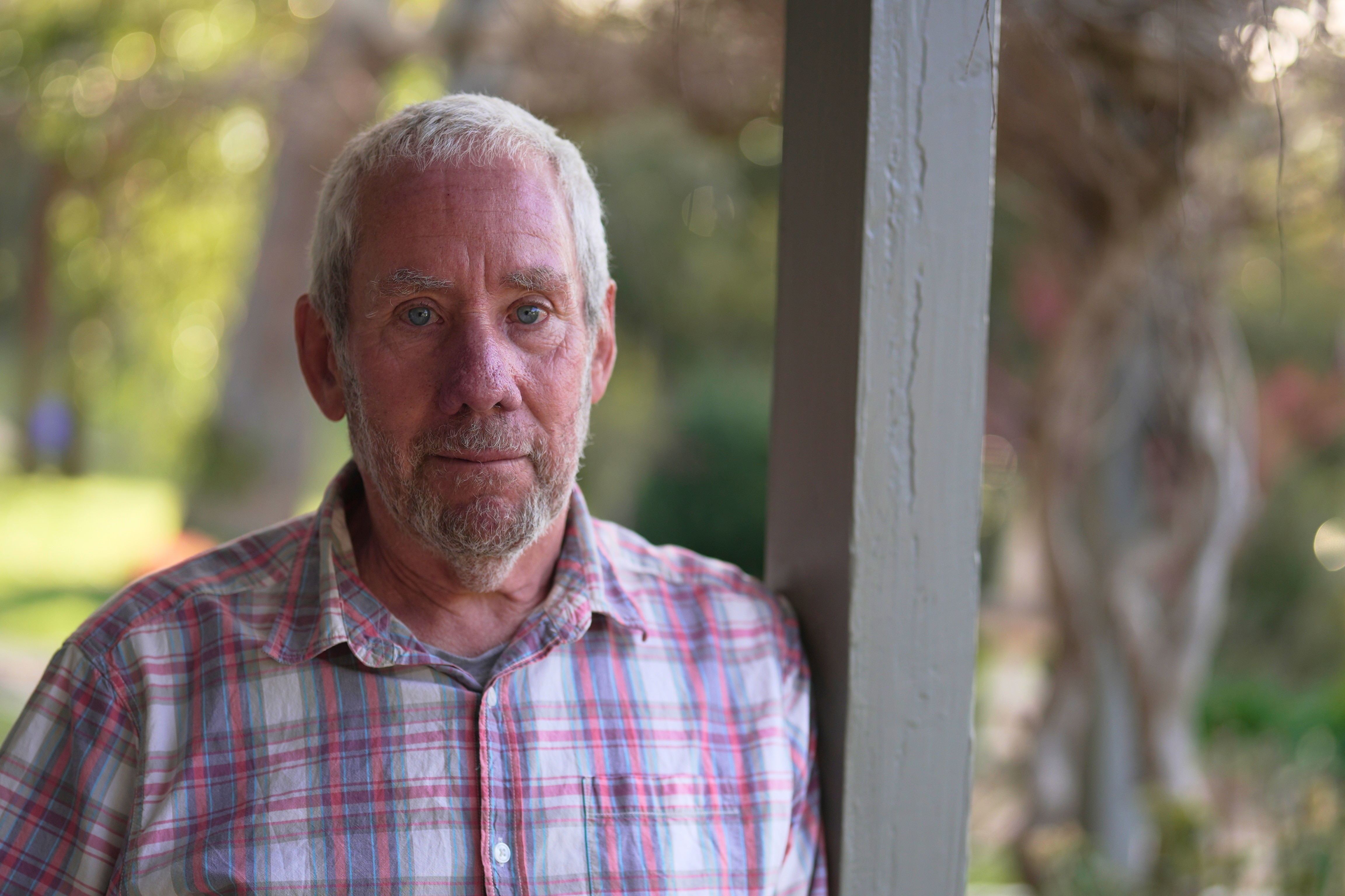 Man with grey hair and light facial hair wears a check shirt and stands outside leaning on a pole.