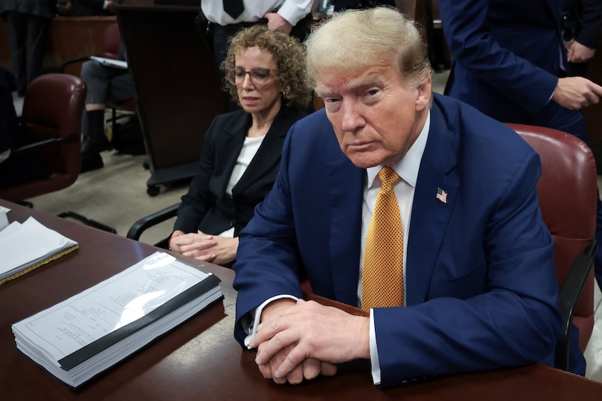 Donald Trump sits in a blue suit and gold tie behind a desk in a courtroom next to a woman