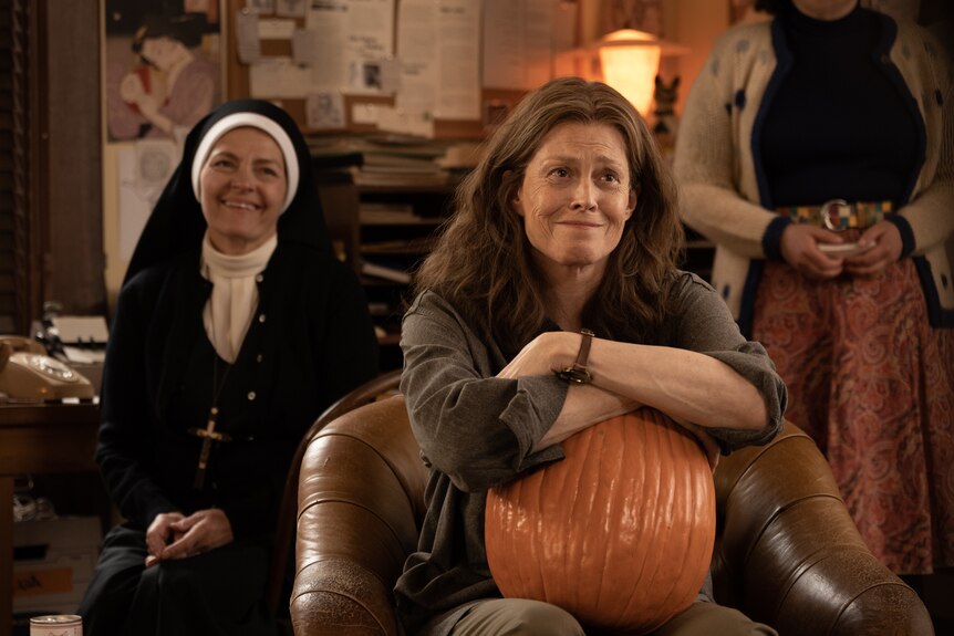 A nun sits smiling behind an older woman with dark hair, who sits with a slight smile, her arms resting on a pumpkin