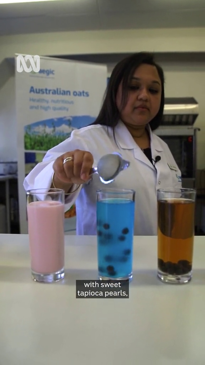 A woman with dark hair and dark-tone skin in a white coat pours something into a glass filled with blue liquid and small balls