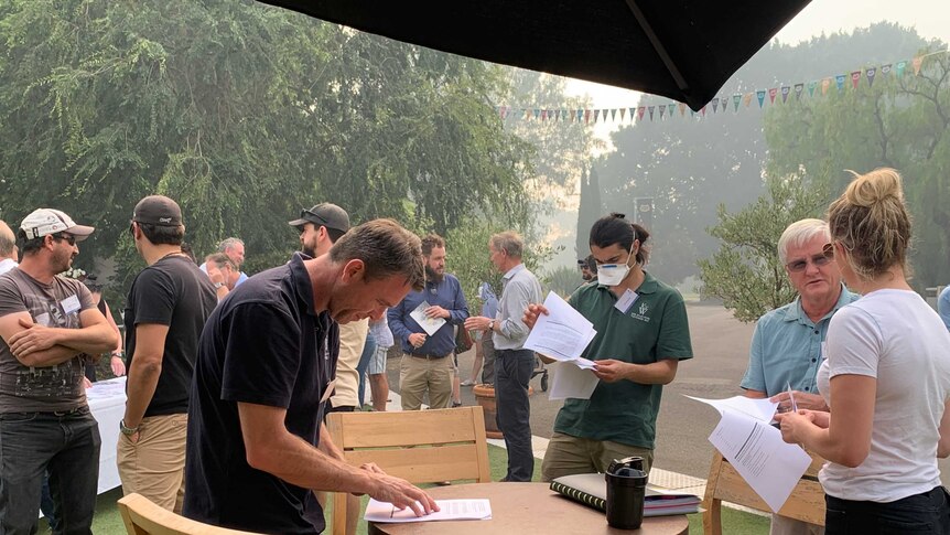 People gather under a smoky sky at a winery and sign paper work. One person wears a smoke mask.