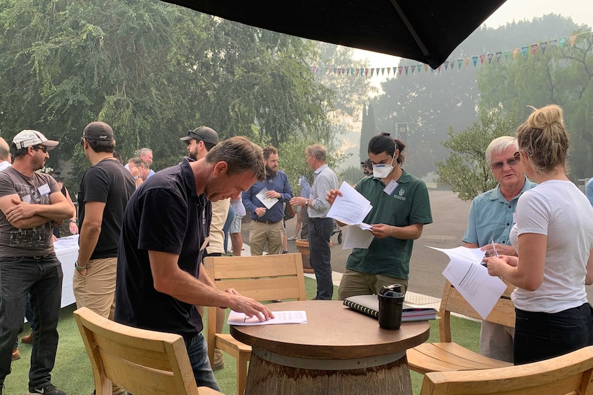 People gather under a smoky sky at a winery and sign paper work. One person wears a smoke mask.