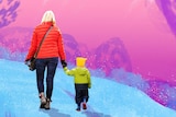 A woman in a red jacket holds the hand of a small child in a green jacket with a yellow beanie on as they walk away