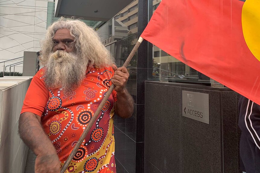 An older Aboriginal man with long grey hair and a beard stands outside court holding an Aboriginal flag and wearing a red shirt.