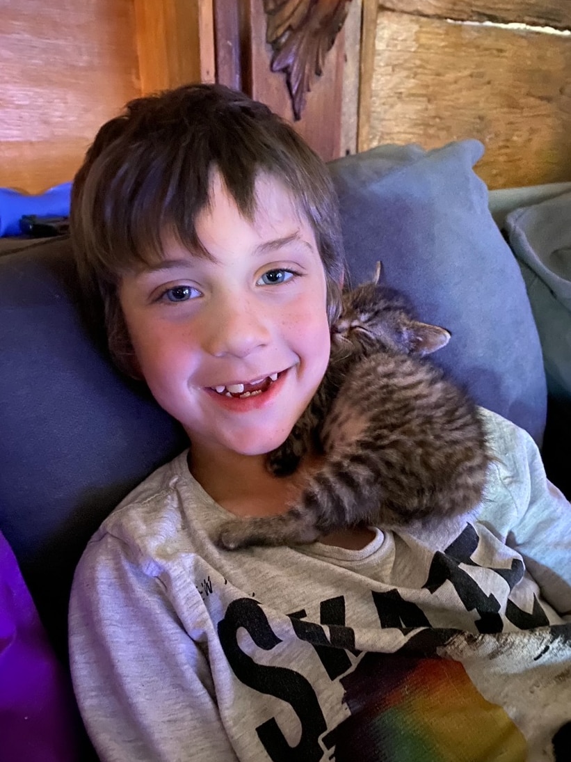 boy with brown hair and missing tooth smiles as he cuddles a sleeping kitten on the couch.