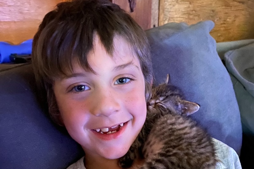 boy with brown hair and missing tooth smiles as he cuddles a sleeping kitten on the couch.