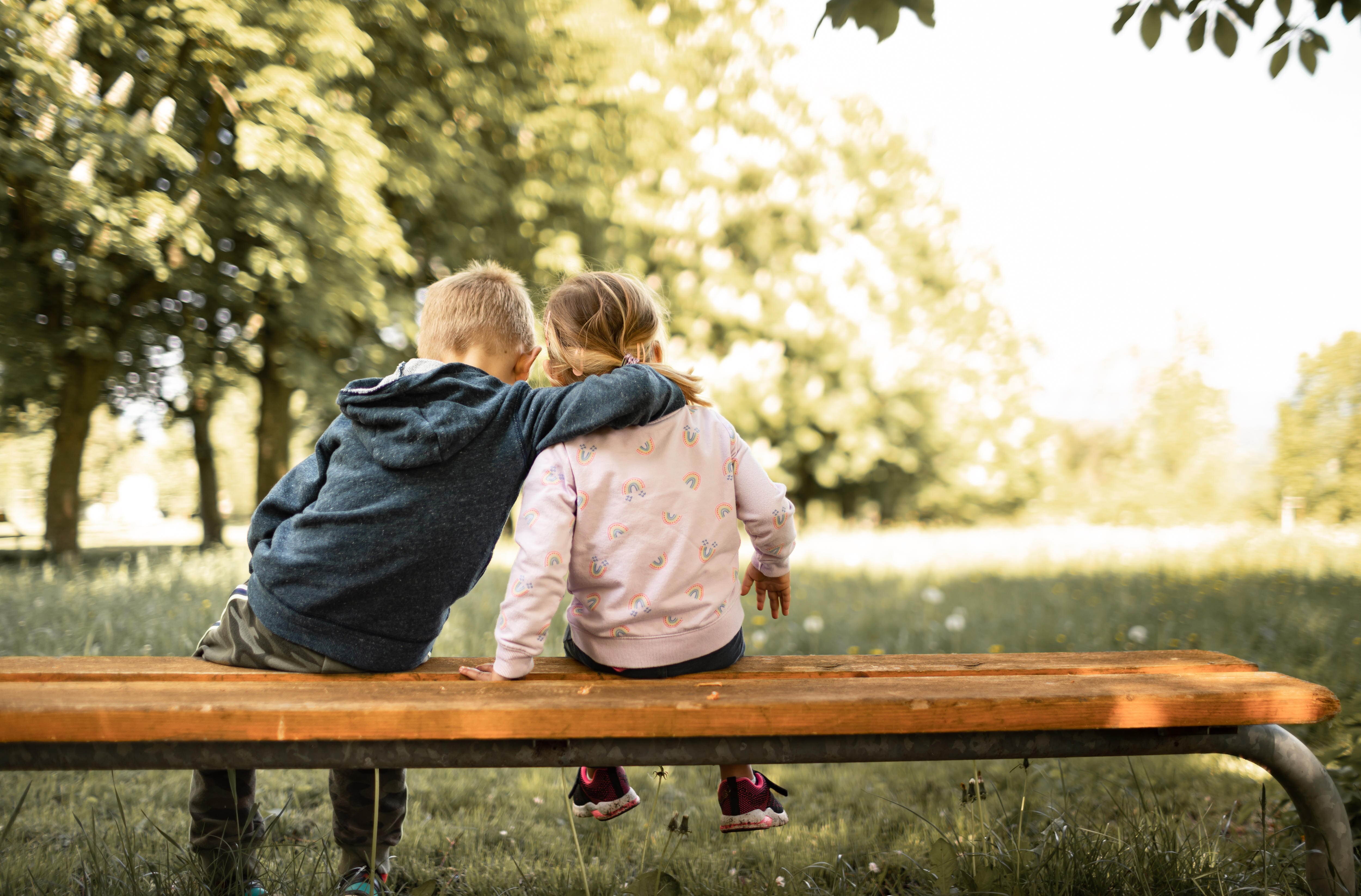 A young boy and girl sit on a bench in the park hugging each other, facing away from the camera.