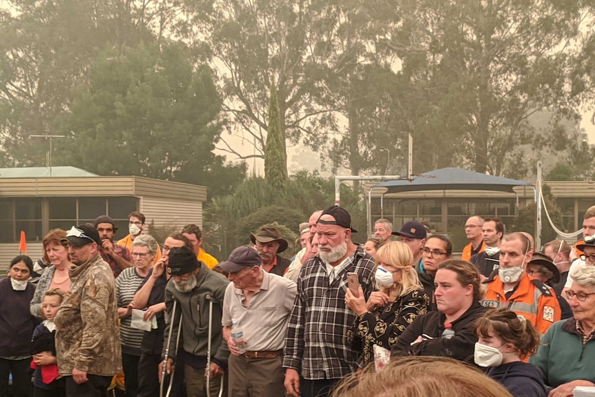 Residents stand listening to a briefing, some are wearing masks. The air is hazy with smoke.