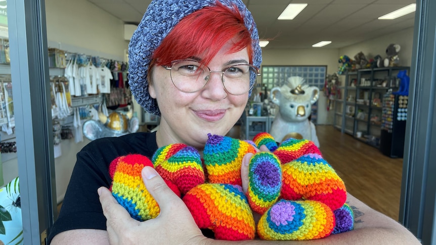 A woman with red hair and wearing a hat stands holding a basket filled with rainbow coloured crocheted hearts.