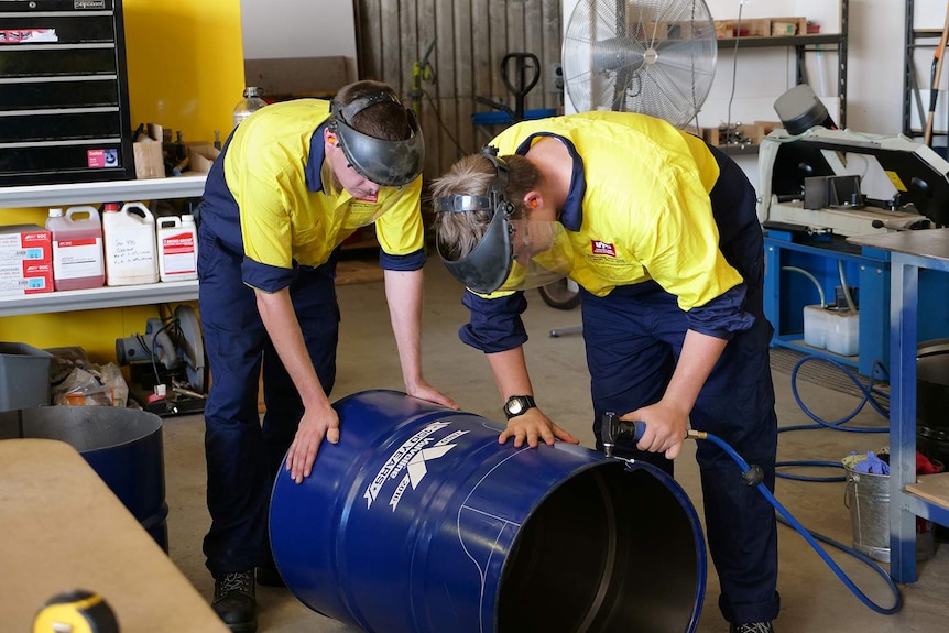 Two men in tradesmen outfits cut the top off an oil drum using an electronic tool.