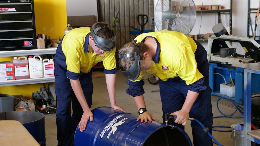 Two men in tradesmen outfits cut the top off an oil drum using an electronic tool.