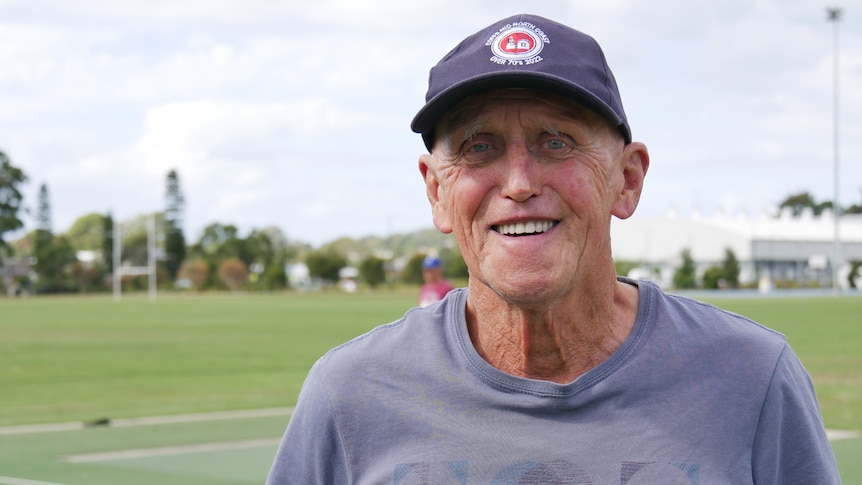 A man in a grey shirt and hat smiles at the camera, while standing on a cricket pitch.
