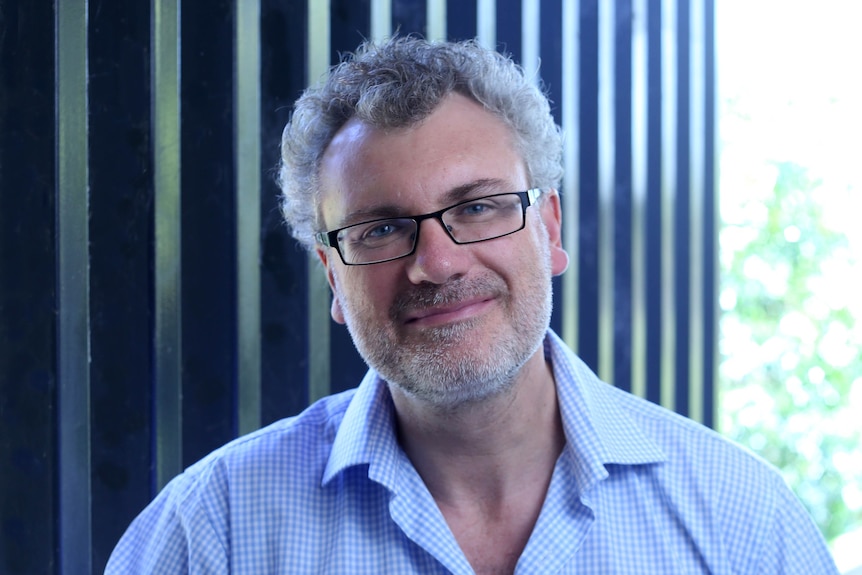Man with curly gray hair and glasses, wearing a collared shirt and standing outside, stares into the camera.