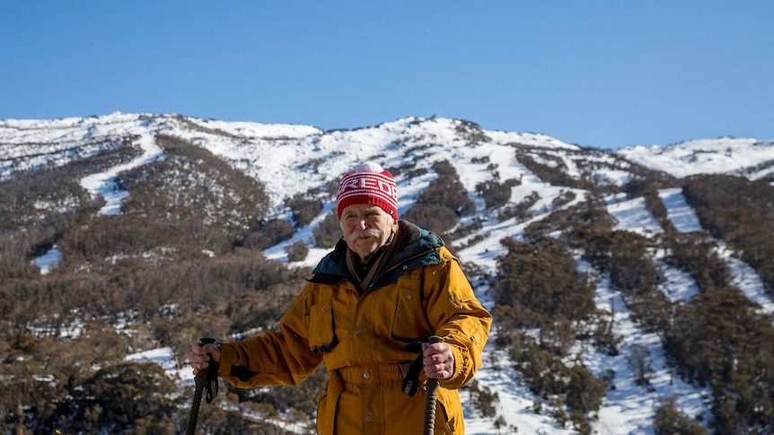 An elderly man in a red beanie and yellow puffy jacket stands on a snow-capped mountain, smiling
