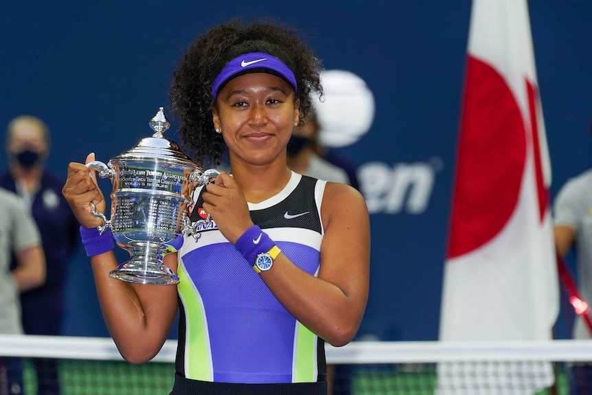 A tennis player smiles as she holds the US Open trophy with a Japanese flag in the background.