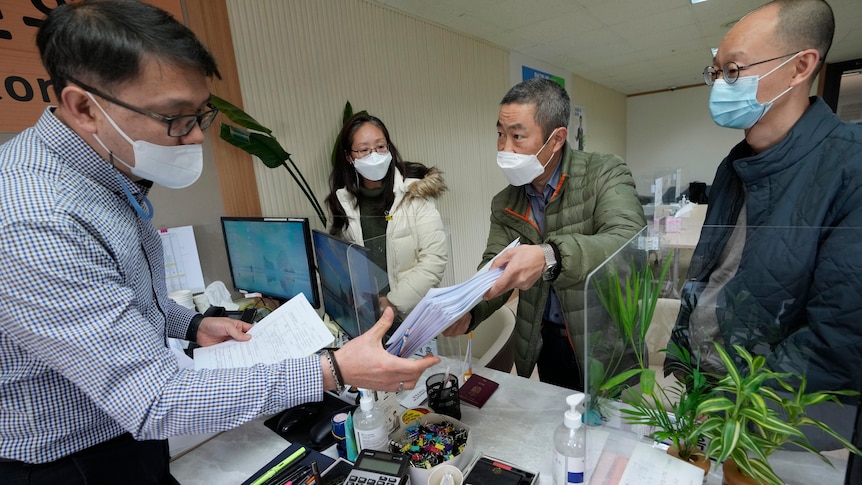 A middle-aged man of Korean descent hands a stack of papers to another man behind a desk in an office building.