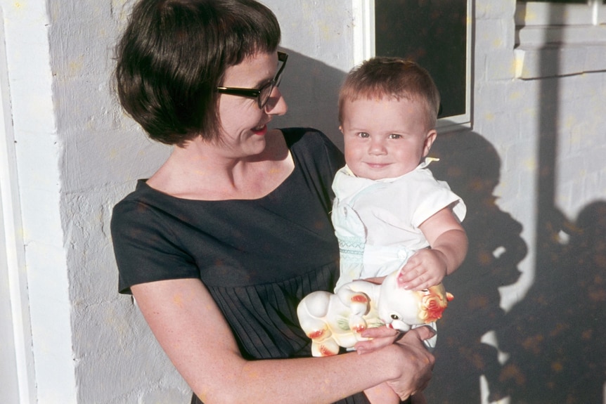 An early 1960s mother holds a baby boy outside the front door of a house