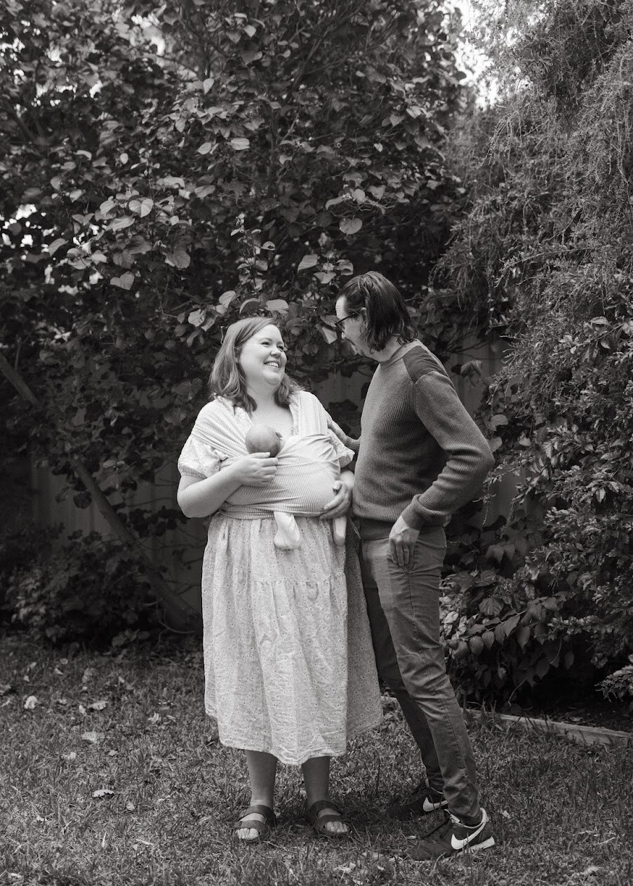 Black and white photo of Chelsea holding her baby with her husband standing by