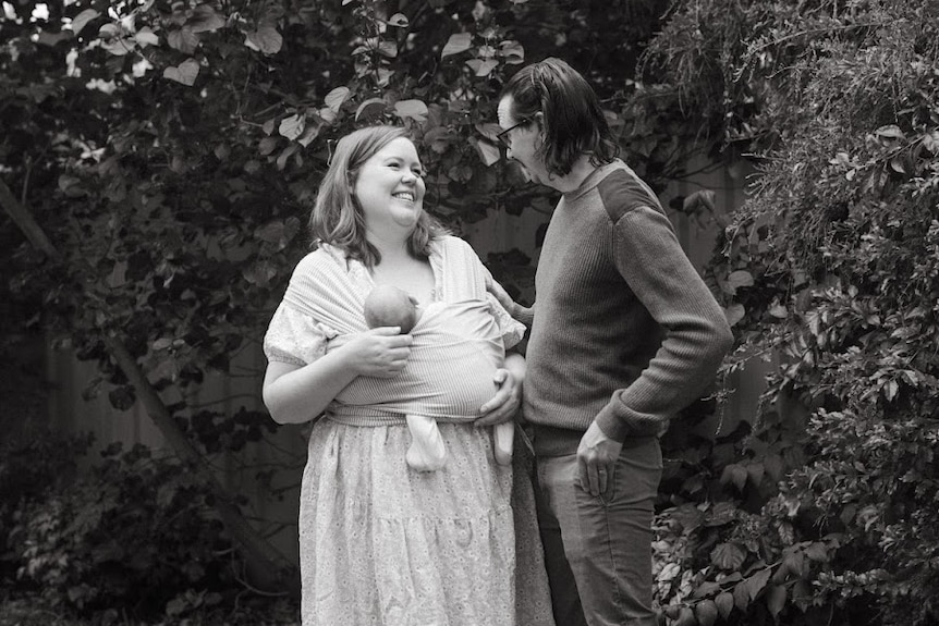 Black and white photo of Chelsea holding her baby with her husband standing by