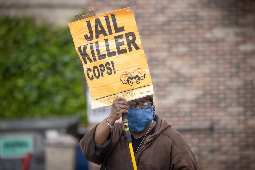 A man wearing a face mask holds a yellow sign saying 'jail killer cops'.