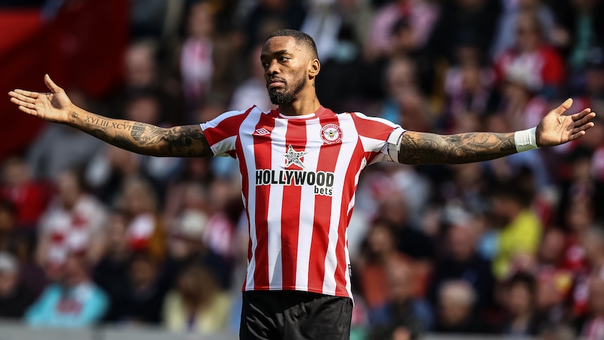 Brentford player Ivan Toney, wearing red and white stripes, stands with his arms outstretched during a Premier League game.