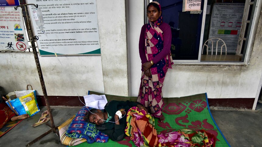 a tea plantation worker lies ona blanket with a drip as his wife stands near him