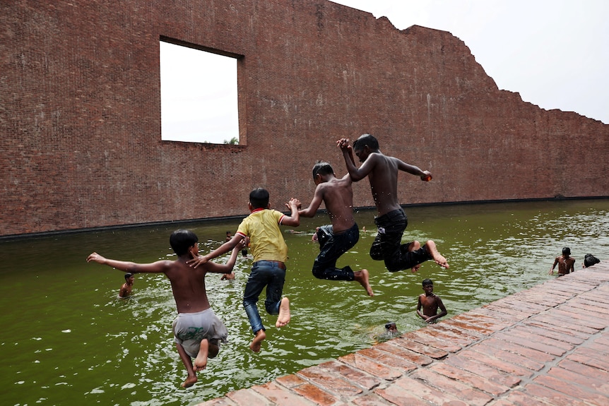 Four boys hold hands and jump into the green water at a large rectangular pool surrounded by brick walls. 