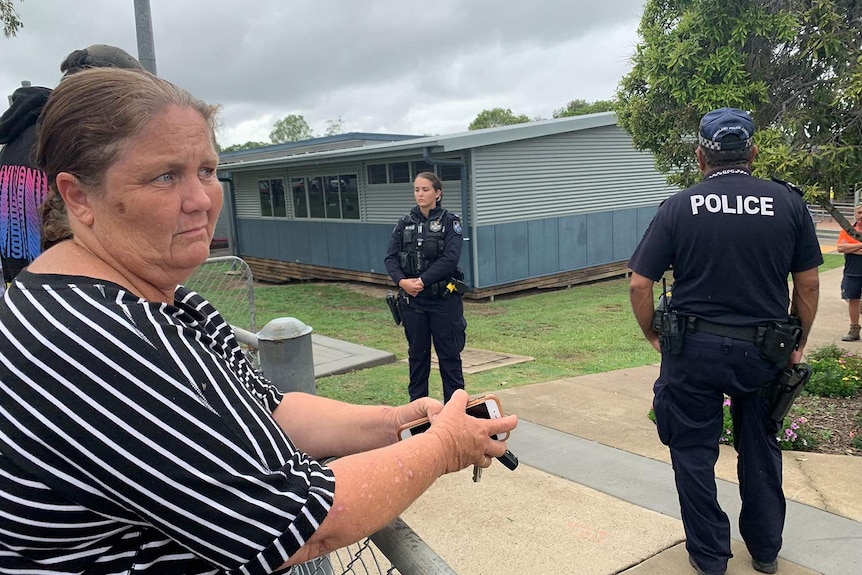 A parent leans on fence outside Rosewood State High School, with two police officers seen on school grounds.