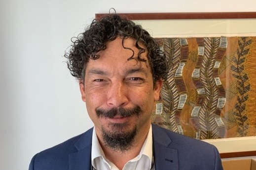 A portrait of a smiling man in his office, in front of an Indigenous artwork, talking about Acknowledgement of Country.