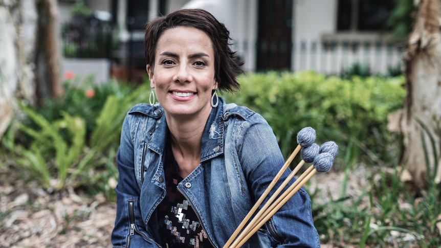 A woman holding sticks used for percussion instruments smiles at the camera. 