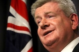 Headshot of Mitch Fifield speaking at a podium with an out of focus Australian flag behind.