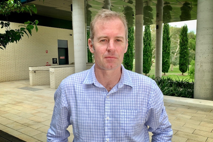 A fair-haired man in his 40s is wearing a pale blue checked shirt as he standing near the undercroft of a university building.