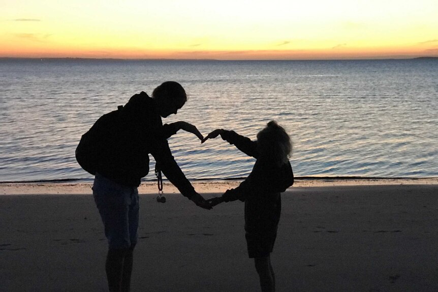 A woman and child on a beach at sunset making a heart with their arms.