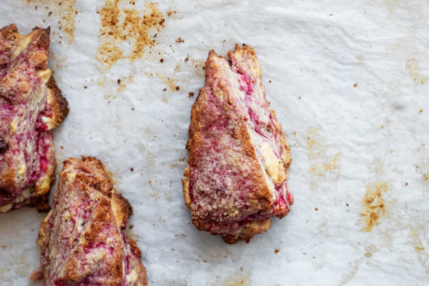 Three flaky triangular scones on a baking sheet with chunks of white chocolate and raspberries.