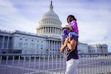A little girl in a face mask sits on a man shoulders as they walk past the US Capitol building which is fenced off