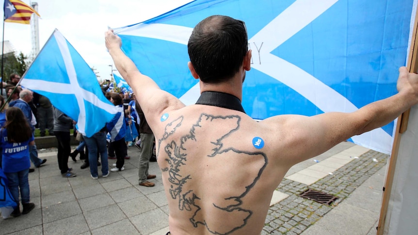 A man with a Scottish map tattooed on his back holds up a Scottish flag.